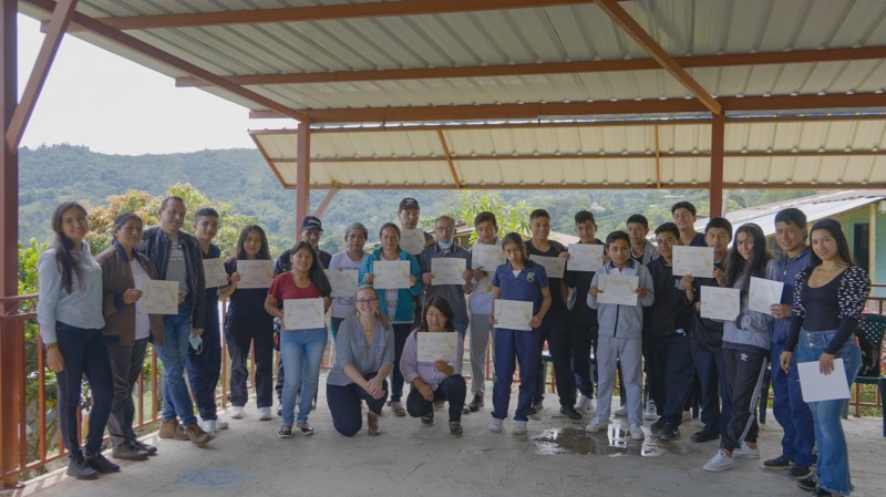A mixed group of around 25 of the Agroecology course participants stand together smiling and holding up their certificates, with the lush forest visible behind